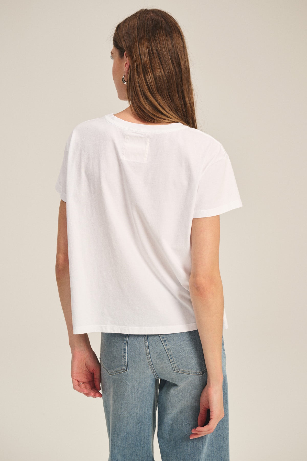 A woman with long brown hair wears the Velvet by Graham & Spencer TIA TEE in white with blue jeans, standing with her back to the camera against a plain background—a timeless essential look.-40305565728961