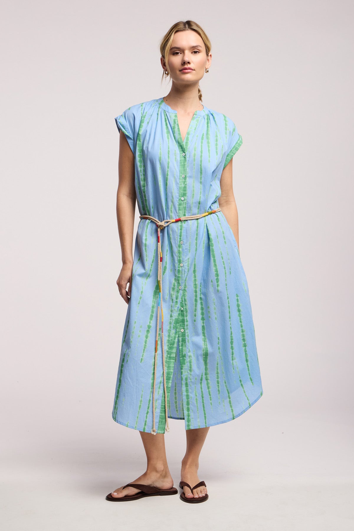A person wears the JADA DRESS by Velvet by Graham & Spencer—a light blue, short-sleeve, button-down midi with green stripes and rope belt—styled with brown sandals for a laid-back California look against a plain backdrop.