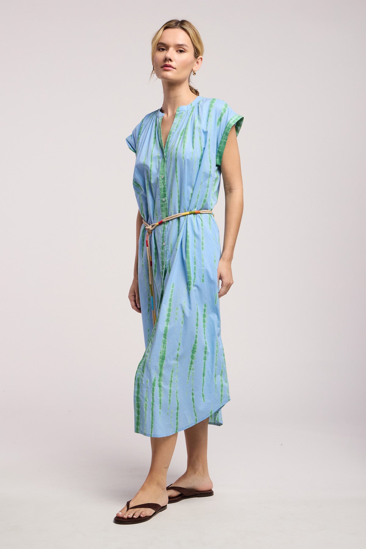 A woman wears the JADA DRESS by Velvet by Graham & Spencer, a light blue California-style knee-length midi with green stripes, short sleeves, a thin waist belt, and black sandals, posed against a plain background.-41645053247681