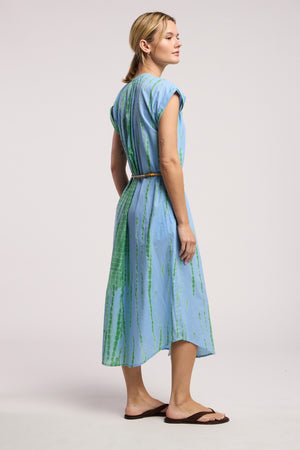 A woman stands sideways in the JADA DRESS by Velvet by Graham & Spencer, a light blue, green-striped midi dress paired with a brown belt and sandals, embodying effortless California style against a plain background.