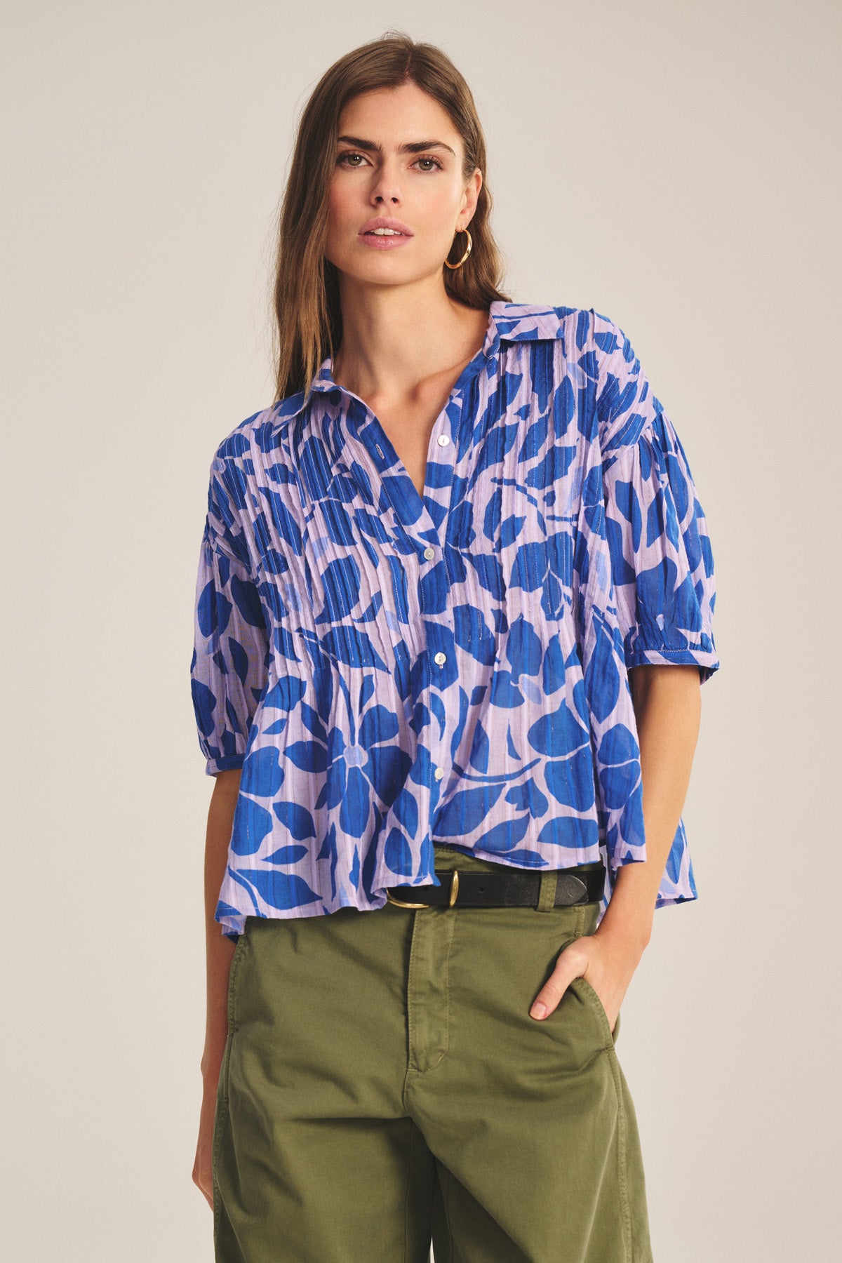 A woman wears the TILDA TOP by Velvet by Graham & Spencer, a blue and lavender patterned cotton short-sleeve top, paired with green high-waisted pleated pants, standing with one hand in her pocket.
