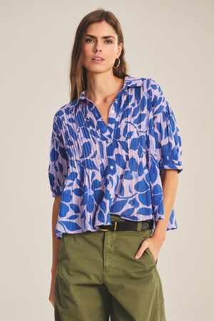 A woman wears the TILDA TOP by Velvet by Graham & Spencer, a blue and lavender patterned cotton short-sleeve top, paired with green high-waisted pleated pants, standing with one hand in her pocket.