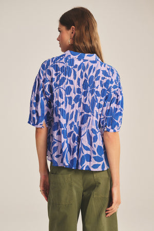 A woman with long brown hair, seen from behind, wears the Velvet by Graham & Spencer TILDA TOP—a blue and white patterned cotton top with half sleeves and pleated details—paired with olive green pants.