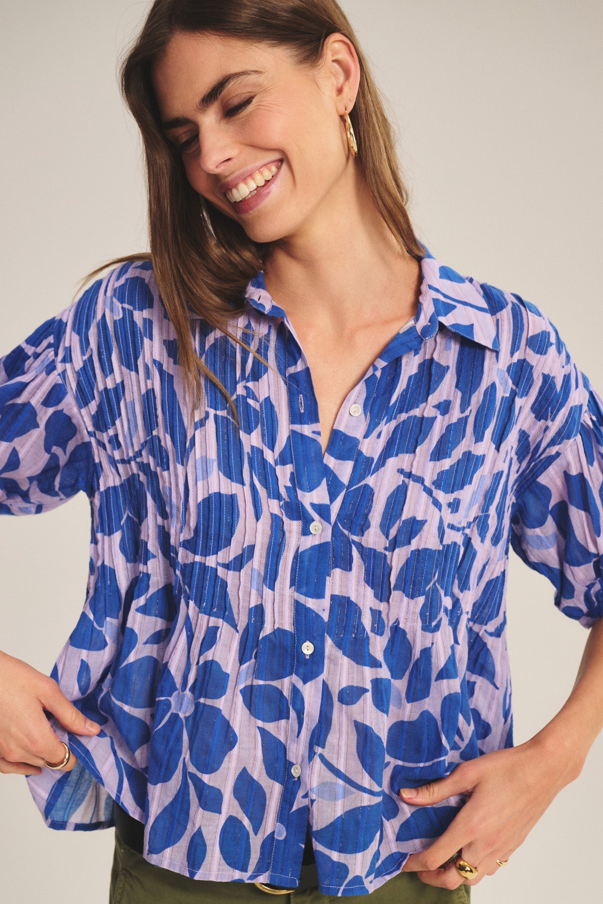 A woman wearing the TILDA TOP by Velvet by Graham & Spencer—a loose, short-sleeve cotton top with a blue floral pattern on light purple and subtle pleats—smiles and looks down.-40808748875969