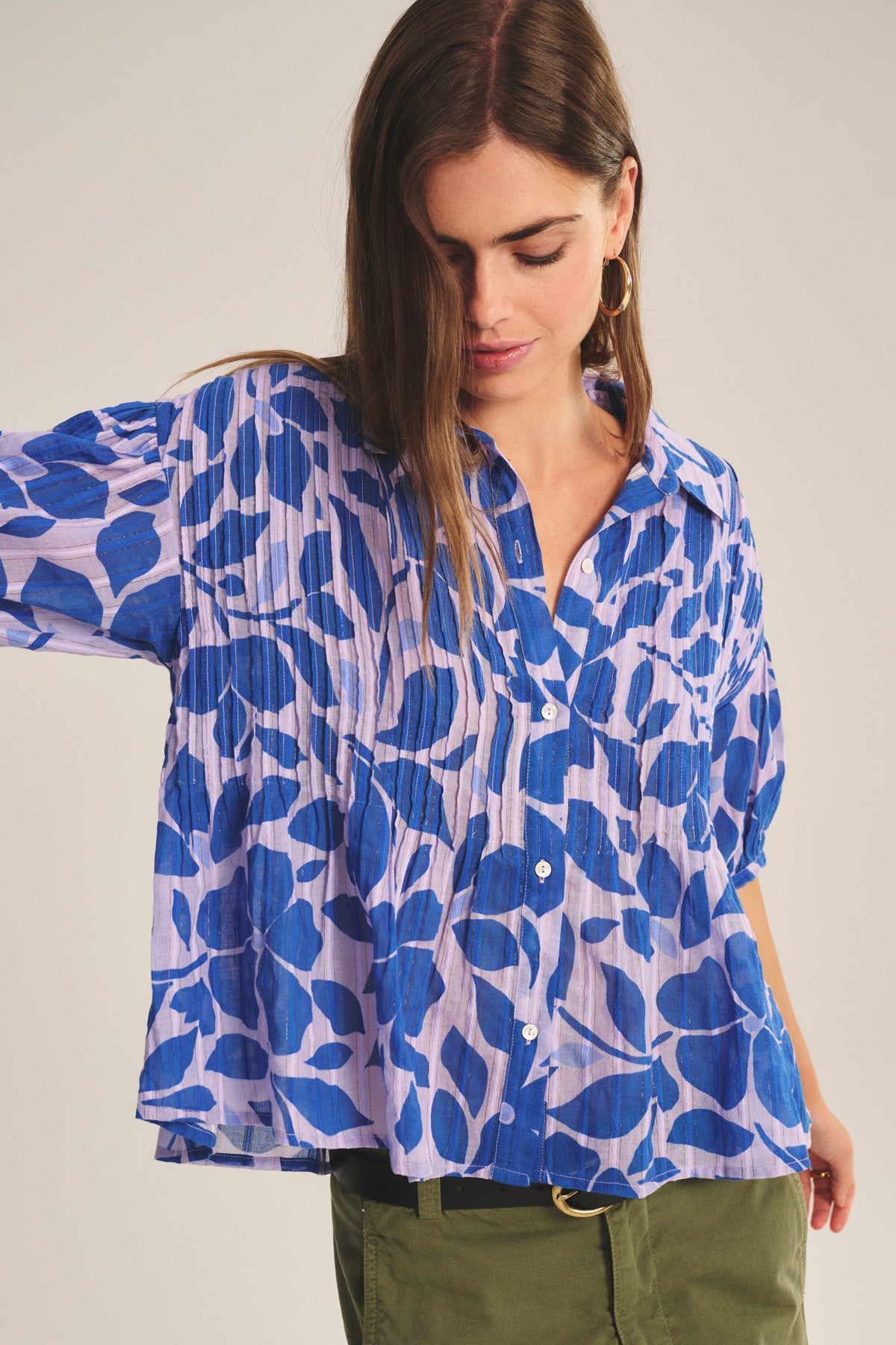 Model wears the TILDA TOP by Velvet by Graham & Spencer—a loose, blue and purple patterned cotton blouse with white buttons and pleated details—paired with olive green pants, looking down with one arm raised.