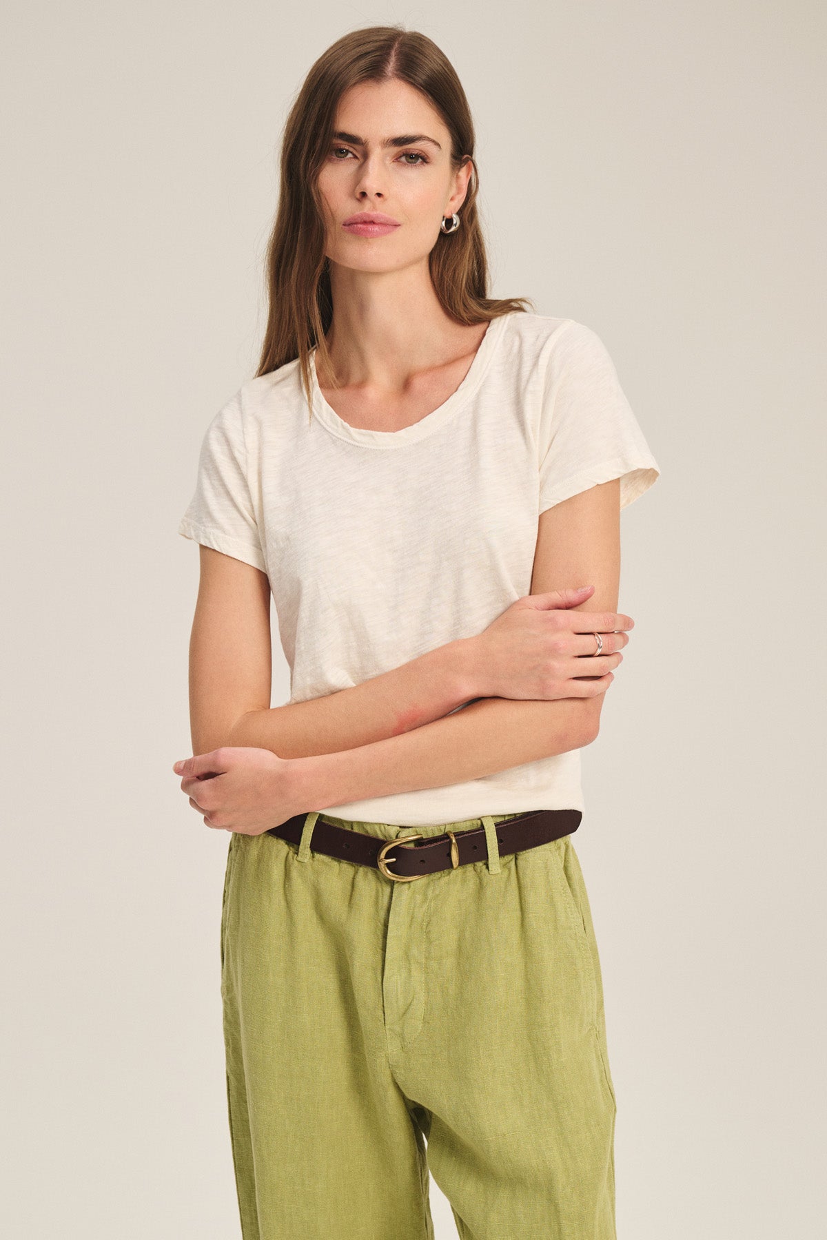 A woman stands against a neutral background, arms crossed, wearing the TILLY TEE by Velvet by Graham & Spencer—white cotton slub with a crew neckline—tucked into light green pants with a dark belt.-41436032139457