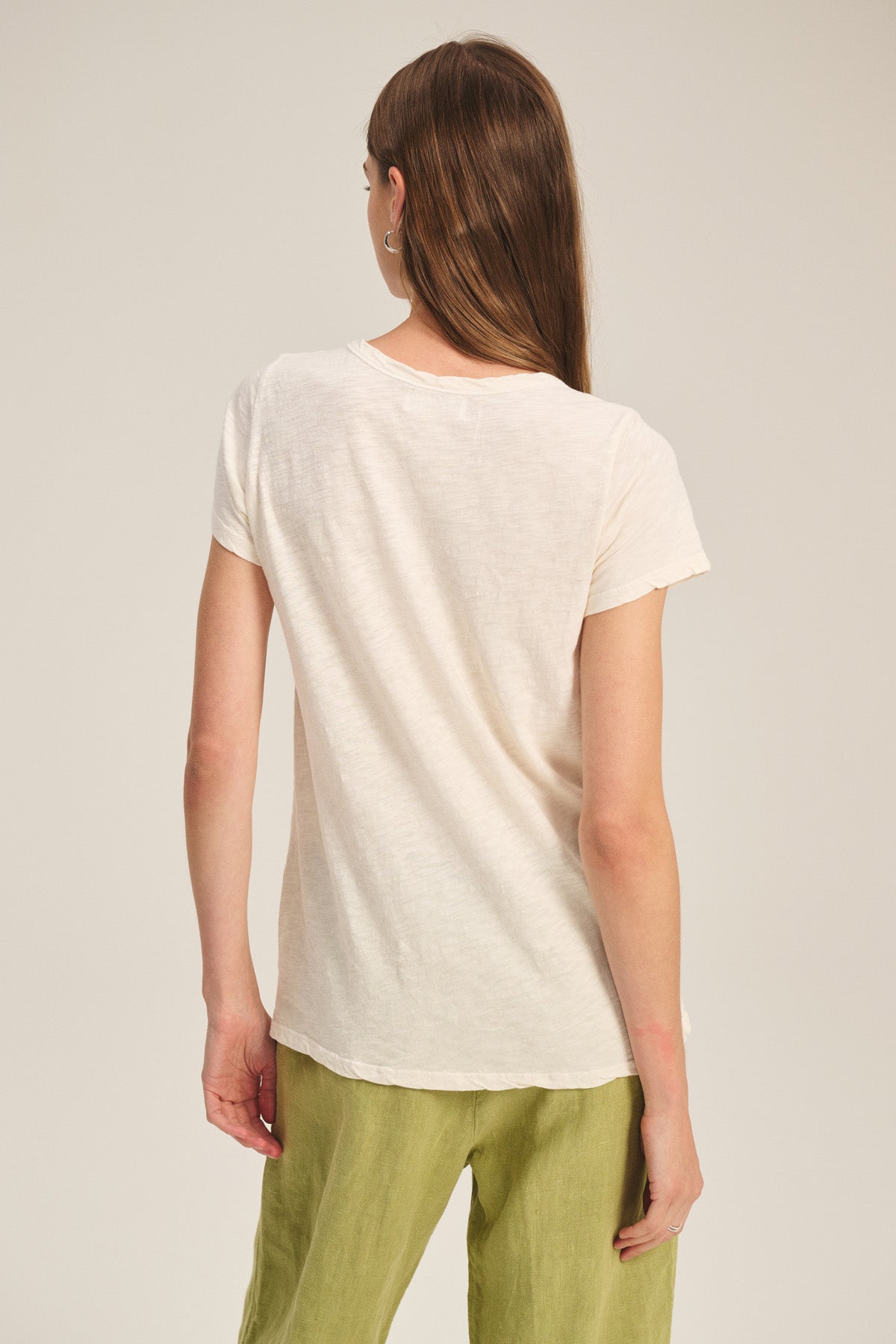A person with long brown hair wears the TILLY TEE by Velvet by Graham & Spencer in a relaxed fit, short sleeve, cotton slub style (off-white), paired with light green pants, standing back to the camera against a plain background.