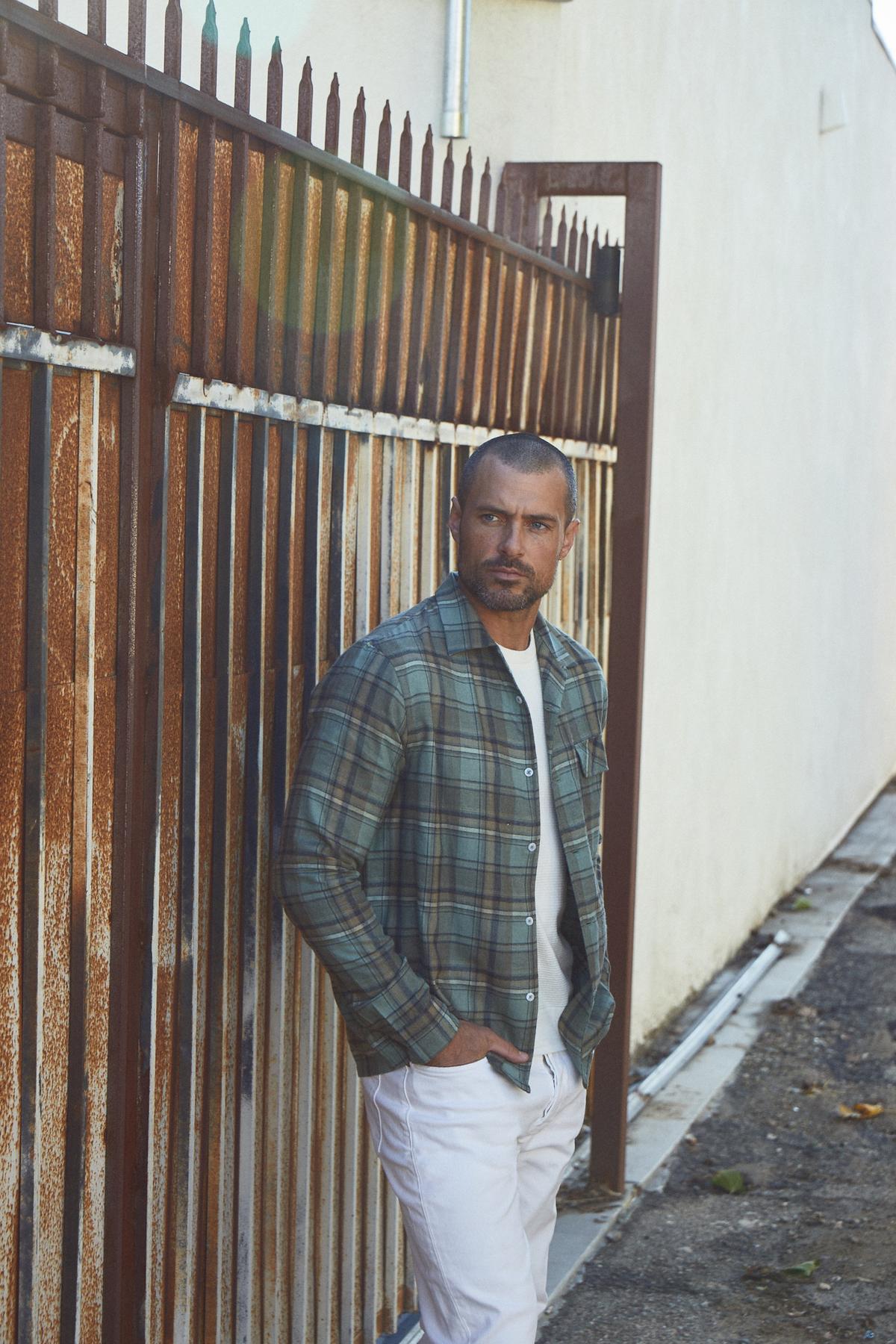   A man in a TIMOTHY PLAID BUTTON-UP SHIRT by Velvet by Graham & Spencer leaning against a fence. 