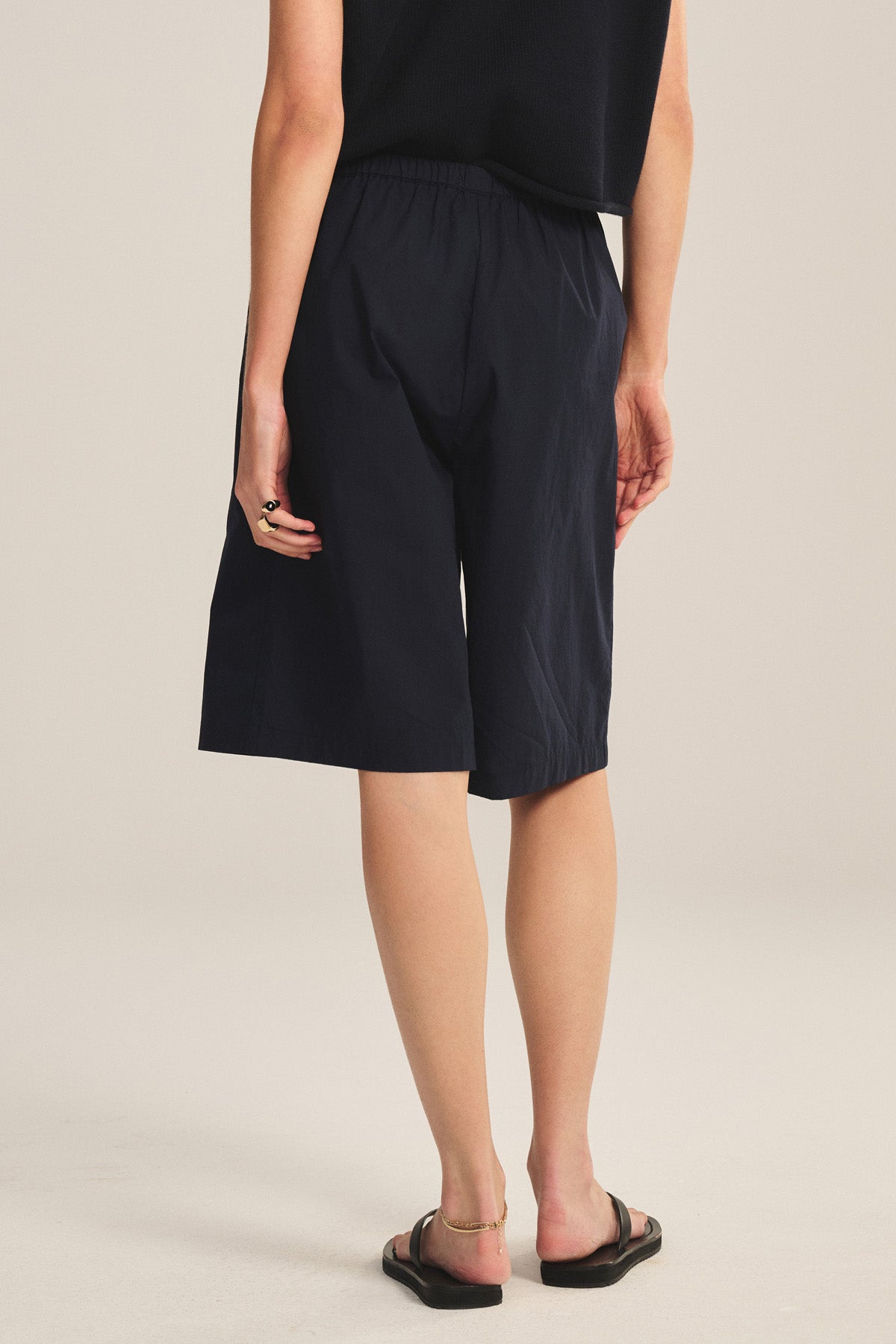 A person stands facing away, wearing a black sleeveless top, Velvet by Graham & Spencer's TOMOKO SHORT pull-on elastic waist cotton poplin shorts with a knee-length silhouette, and black sandals.-41044164378817
