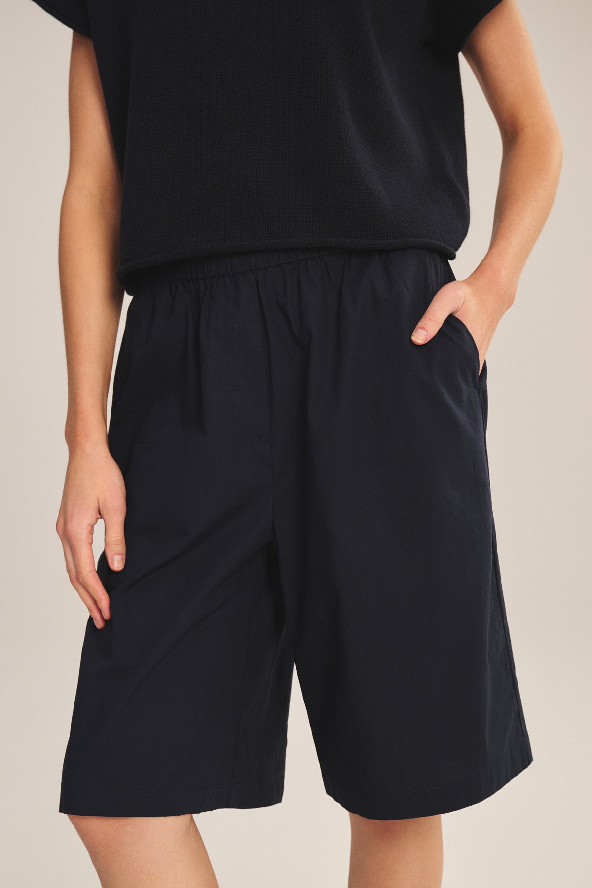 Wearing the TOMOKO SHORT by Velvet by Graham & Spencer—pull-on, knee-length cotton poplin shorts with elastic waist and pockets—paired with a black short-sleeve shirt, model stands against a neutral background.-41044164346049