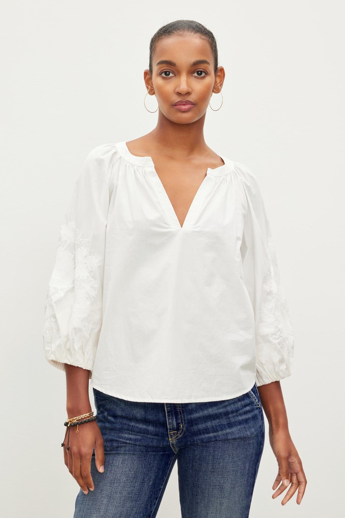   A person wearing a white TRINA EMBROIDERED BOHO TOP by Velvet by Graham & Spencer and blue jeans stands against a plain background. 