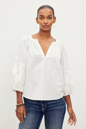 A person wearing a white TRINA EMBROIDERED BOHO TOP by Velvet by Graham & Spencer and blue jeans stands against a plain background.