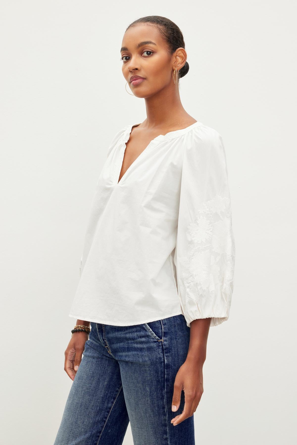   A person stands wearing a white TRINA EMBROIDERED BOHO TOP by Velvet by Graham & Spencer with a deep V-neck and puffy sleeves, paired with blue jeans, against a plain white background. 