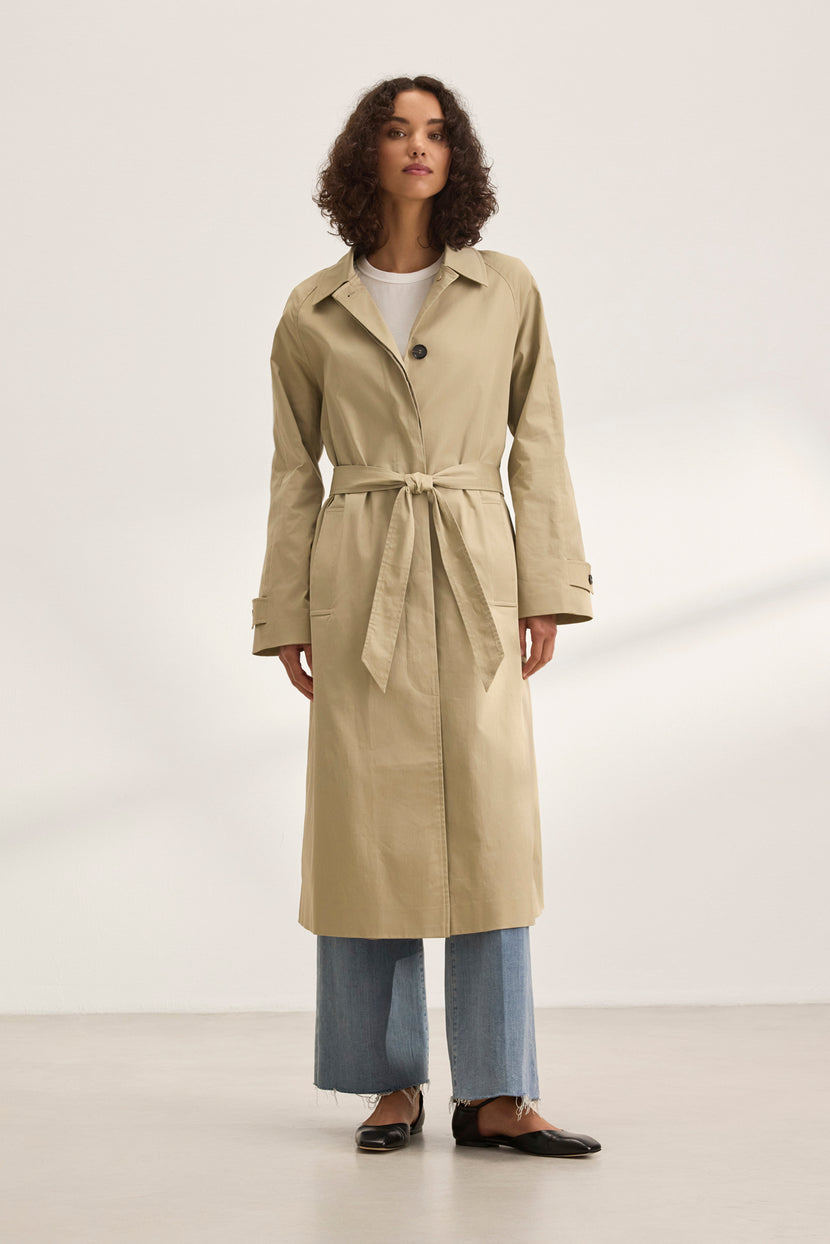 A person models the versatile RAHEAL TRENCH COAT by Velvet by Graham & Spencer. They pair it with light blue jeans and black shoes against a plain backdrop, highlighting the beige cotton-elastane coat's stylish design with a belt.