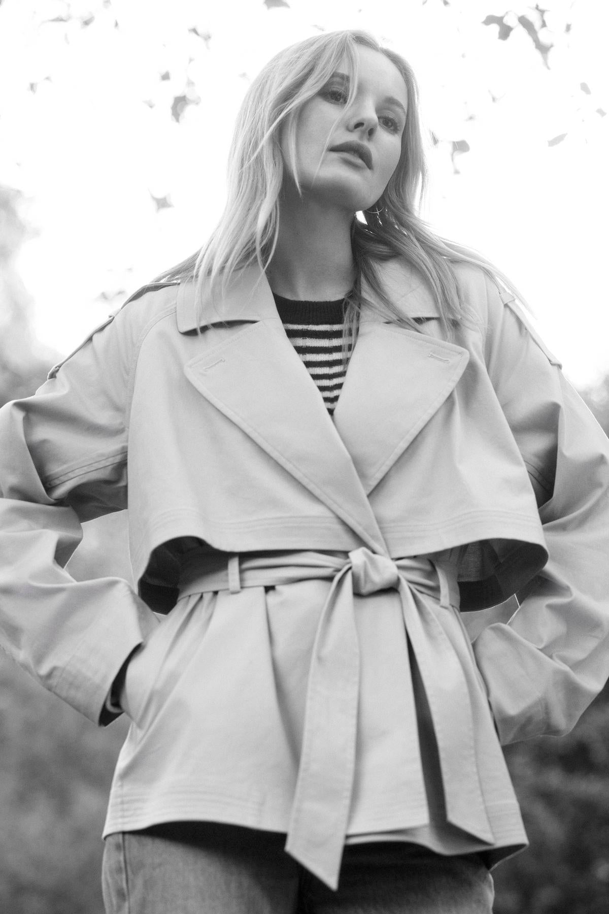   A woman stands outdoors wearing a belted trench coat over the Monica Cashmere Sweater by Velvet by Graham & Spencer, with her hands in her pockets, looking slightly upwards. The photo is in black and white, capturing the essence of timeless wardrobe essentials. 