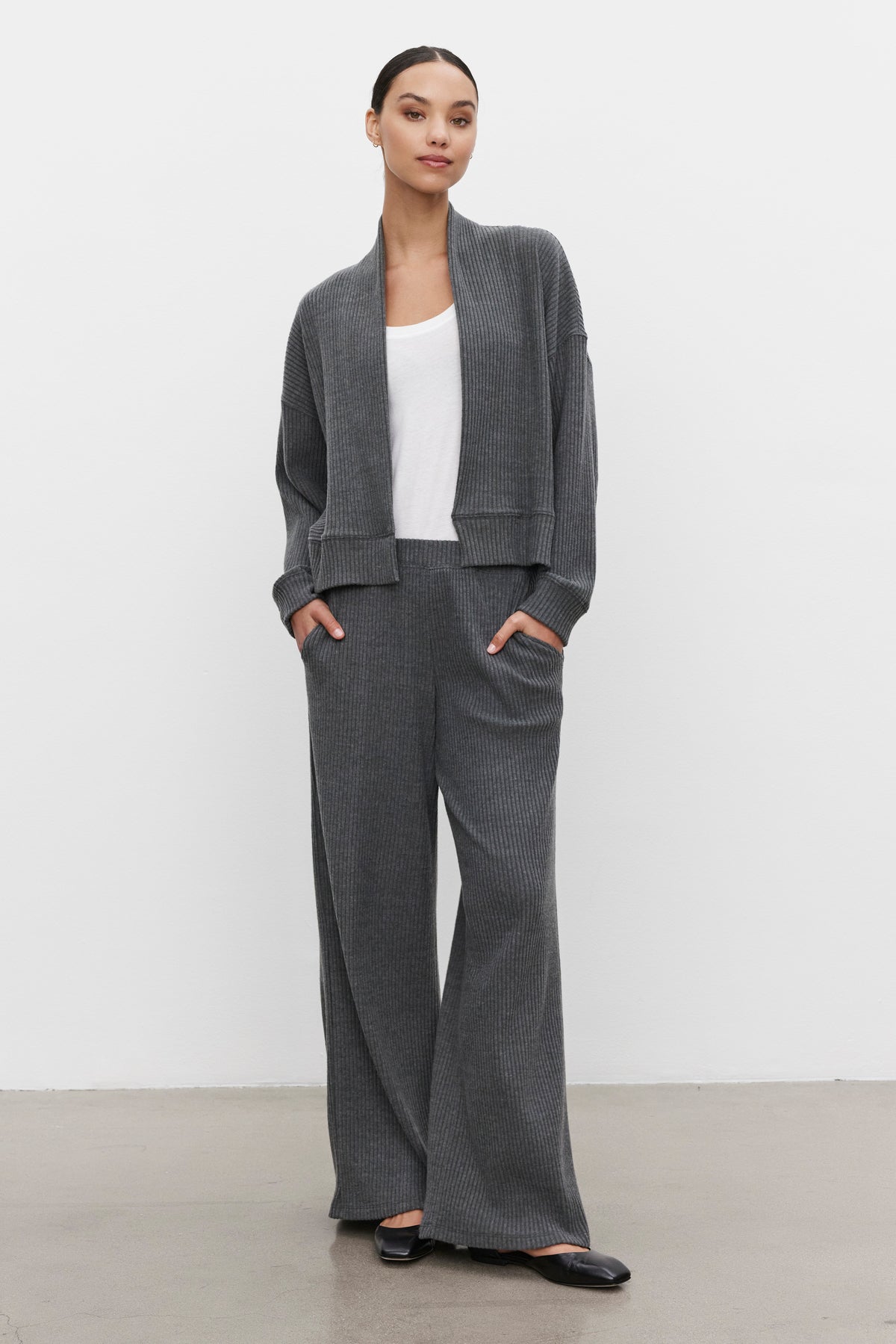   A person stands against a white background wearing the ultra-soft and cozy LYNNETTE RIBBED CARDIGAN in gray by Velvet by Graham & Spencer, paired with matching wide-leg pants. Their hands are in the pockets, completing the look with a white top and black shoes. 