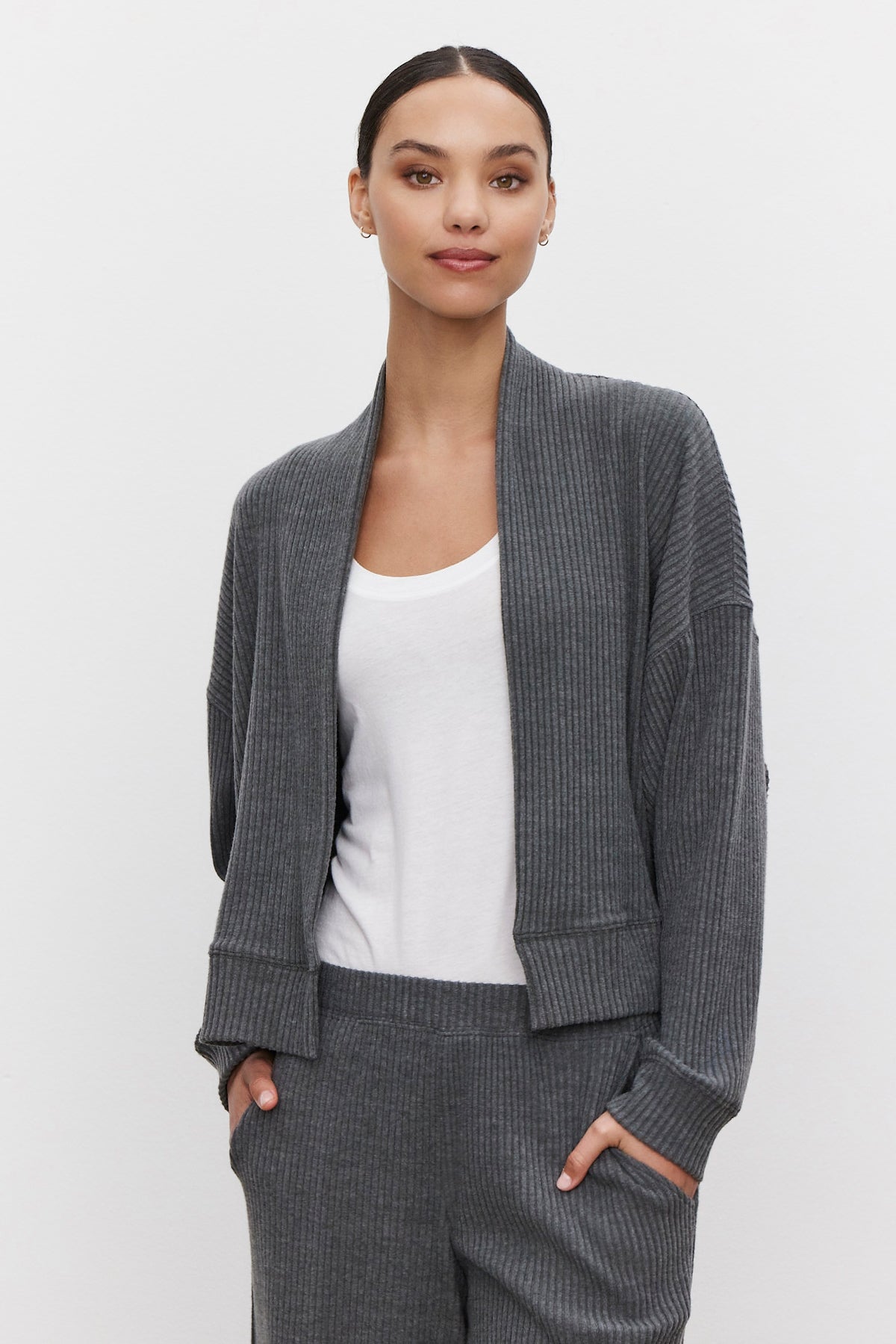   A person wearing an ultra-soft and cozy Velvet by Graham & Spencer LYNNETTE RIBBED CARDIGAN in grey over a white top with matching grey ribbed pants, standing against a plain light background. 