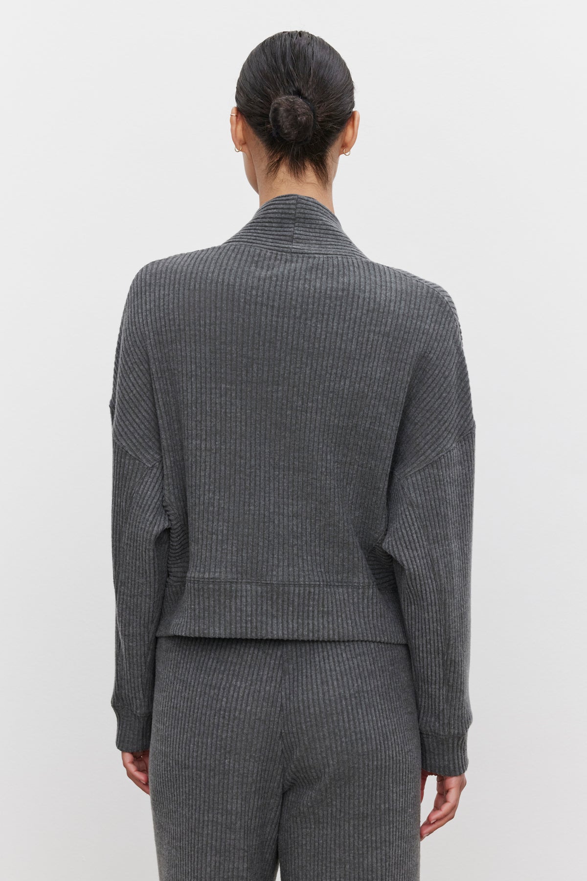   A person with tied back hair stands facing away, dressed in the ultra-soft and cozy LYNNETTE RIBBED CARDIGAN and matching pants from Velvet by Graham & Spencer. The background is plain and white. 