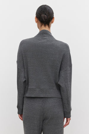 A person with tied back hair stands facing away, dressed in the ultra-soft and cozy LYNNETTE RIBBED CARDIGAN and matching pants from Velvet by Graham & Spencer. The background is plain and white.