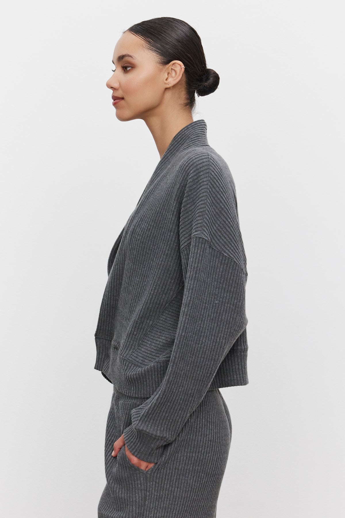   A woman in profile view wearing an ultra-soft and cozy LYNNETTE RIBBED CARDIGAN by Velvet by Graham & Spencer, along with matching pants, stands against a plain white background. Her dark hair is pulled back into a sleek bun. 