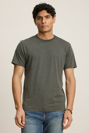 A man with short curly hair wears the ZANDER TEE by Velvet by Graham & Spencer, a relaxed fit dark gray t-shirt with a ribbed crew neckline, paired with blue jeans against a neutral background.