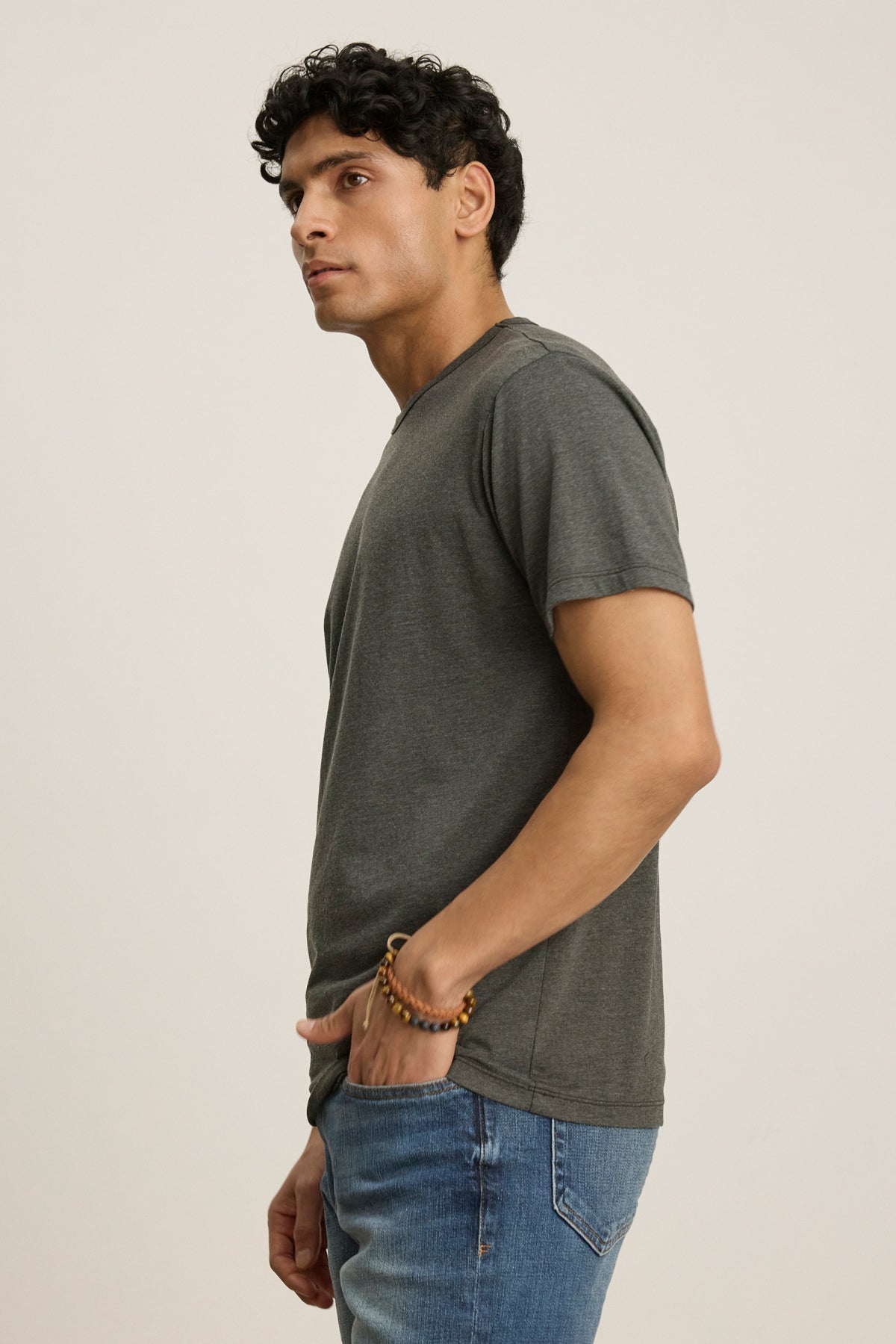A man stands sideways with one hand in his pocket, wearing the Velvet by Graham & Spencer ZANDER TEE in grey—a relaxed fit, short-sleeve cotton-modal shirt—paired with blue jeans against a plain background.-39446054961345