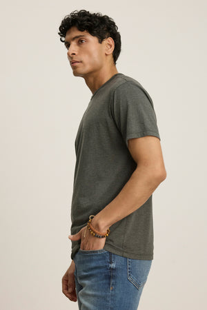 A man stands sideways with one hand in his pocket, wearing the Velvet by Graham & Spencer ZANDER TEE in grey—a relaxed fit, short-sleeve cotton-modal shirt—paired with blue jeans against a plain background.