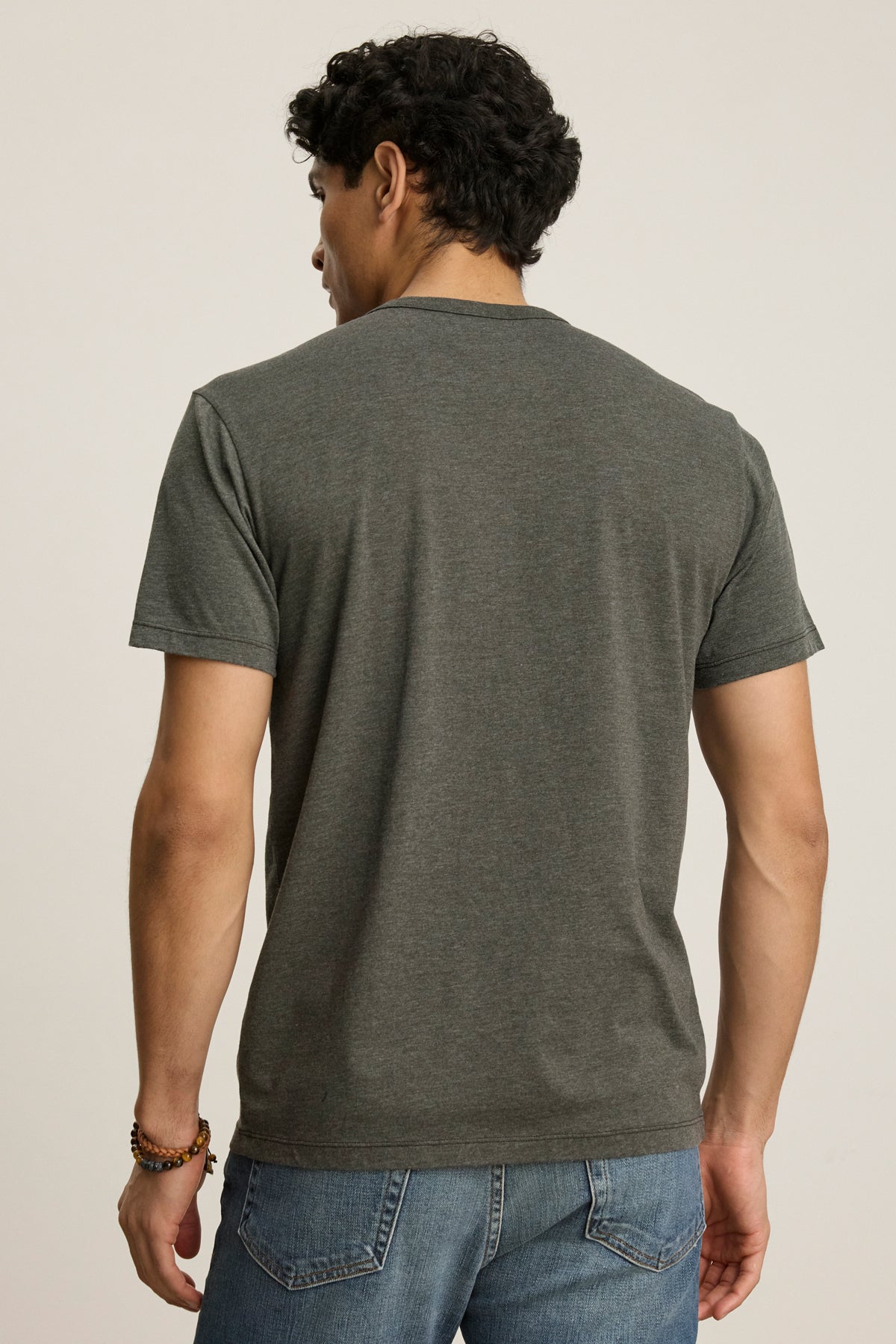 A man with short, curly dark hair is shown from the back wearing the Velvet by Graham & Spencer ZANDER TEE in heather gray and blue jeans, standing against a plain background.-39446054994113