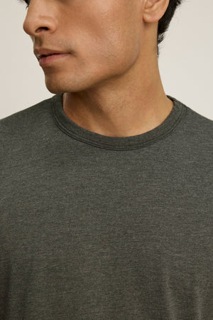 A close-up of a person wearing the ZANDER TEE by Velvet by Graham & Spencer—a plain dark gray, relaxed-fit t-shirt with a ribbed crew neckline—showing the lower half of their face and upper torso.