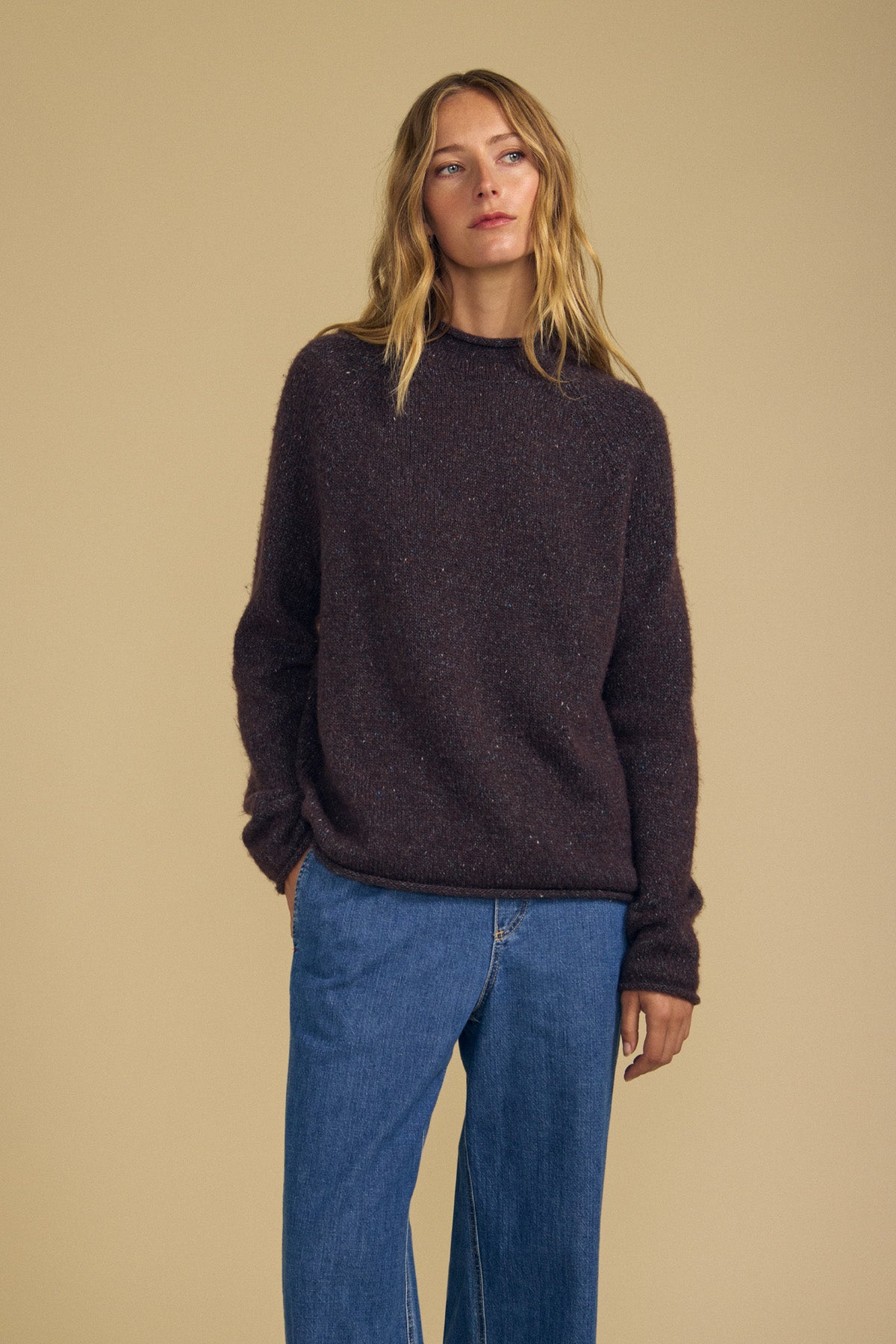 Wearing the JOELLE SWEATER by Velvet by Graham & Spencer, a person with long blonde hair stands against a plain beige background with one hand in their pocket, pairing the dark brown wool blend sweater with blue jeans.-42063767568577