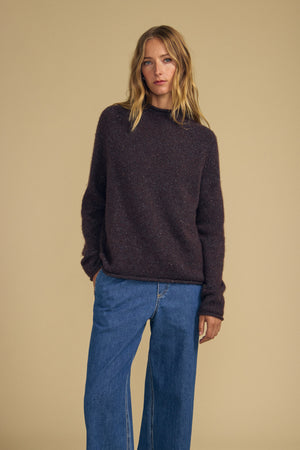 A person with long blonde hair wears the JOELLE SWEATER by Velvet by Graham & Spencer, a dark brown mock neck sweater, paired with blue wide-leg jeans and stands against a plain beige background.