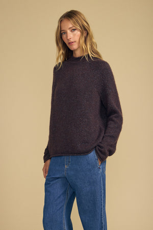A woman with long blonde hair wears the JOELLE SWEATER by Velvet by Graham & Spencer—a loose dark brown mock neck style—paired with blue jeans, standing with her hands in her pockets against a plain beige background.