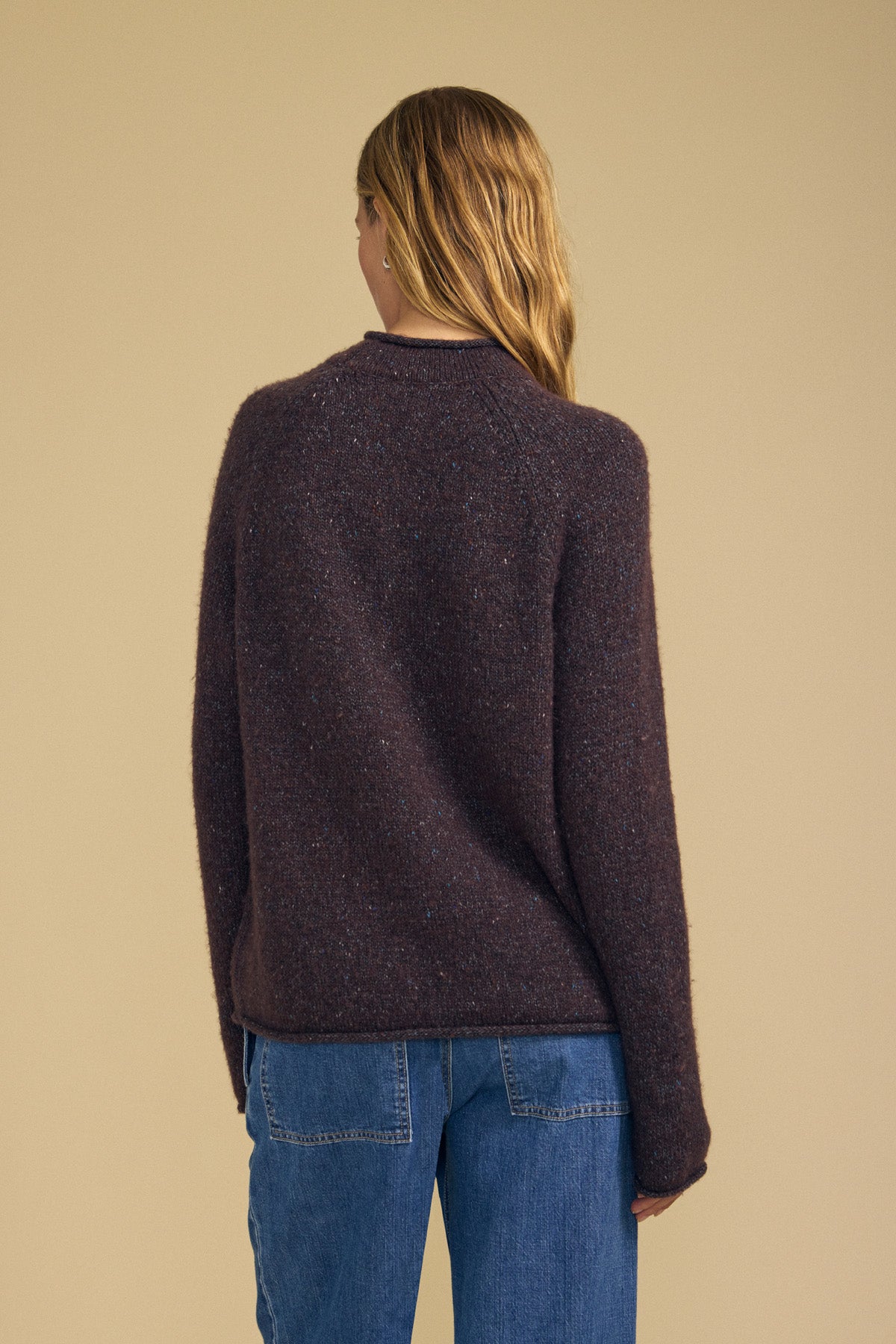 A person with long hair is seen from behind, standing against a plain beige background, wearing the Velvet by Graham & Spencer JOELLE SWEATER in dark brown paired with blue jeans.-42063767666881