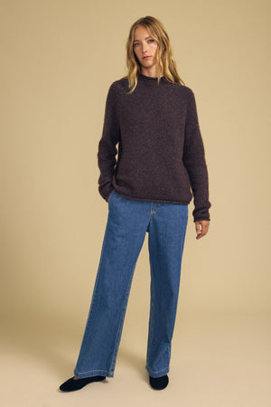 A person stands against a beige background wearing the JOELLE SWEATER by Velvet by Graham & Spencer, paired with wide-leg blue jeans and black shoes, hands in pockets.