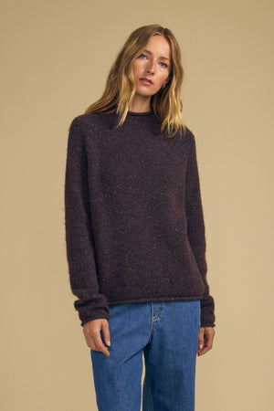 A person with long, blonde hair wears the JOELLE SWEATER by Velvet by Graham & Spencer—a dark brown wool blend piece—paired with blue jeans, standing against a plain beige background.