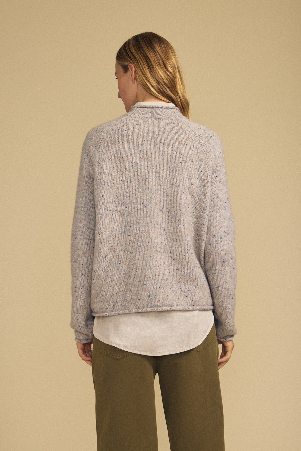 A person with long hair is seen from behind, standing against a beige background and wearing the JOELLE SWEATER by Velvet by Graham & Spencer—a light gray speckled tweed knit—layered over a white shirt and paired with olive pants.-42063767470273