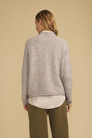A person with long hair is seen from behind, standing against a beige background and wearing the JOELLE SWEATER by Velvet by Graham & Spencer—a light gray speckled tweed knit—layered over a white shirt and paired with olive pants.