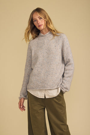 Wearing the JOELLE SWEATER by Velvet by Graham & Spencer, a person stands against a beige background, layering the light gray wool blend sweater over a collared shirt and pairing it with olive green pants.