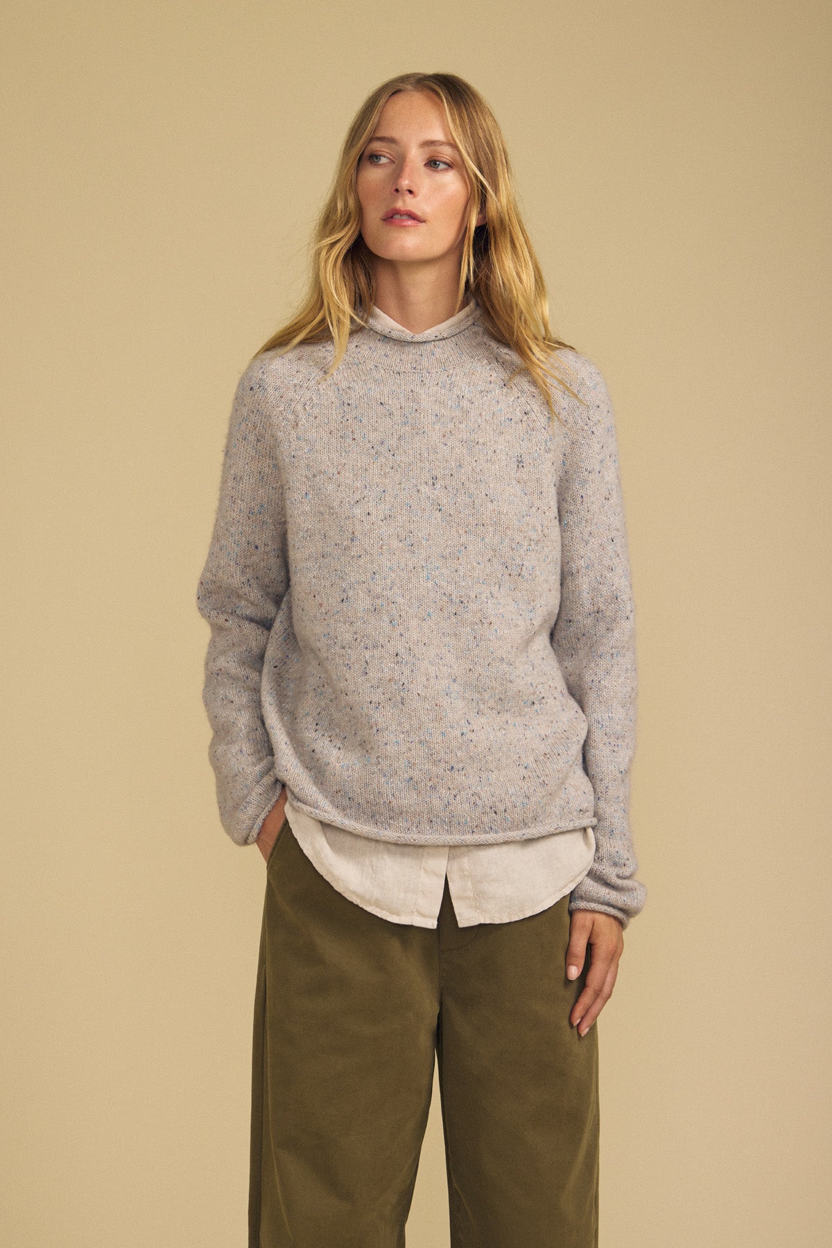 A woman stands against a beige background wearing the JOELLE SWEATER by Velvet by Graham & Spencer—a loose, light gray wool blend layered over a collared shirt—and olive green wide-leg pants, with her hands in her pockets and a neutral expression.-42063767765185