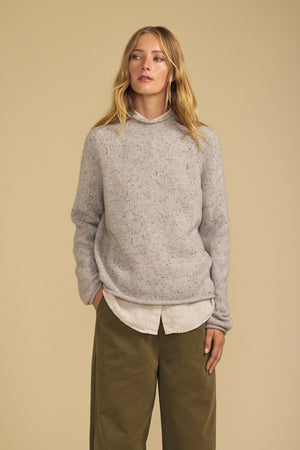 A woman stands against a beige background wearing the JOELLE SWEATER by Velvet by Graham & Spencer—a loose, light gray wool blend layered over a collared shirt—and olive green wide-leg pants, with her hands in her pockets and a neutral expression.