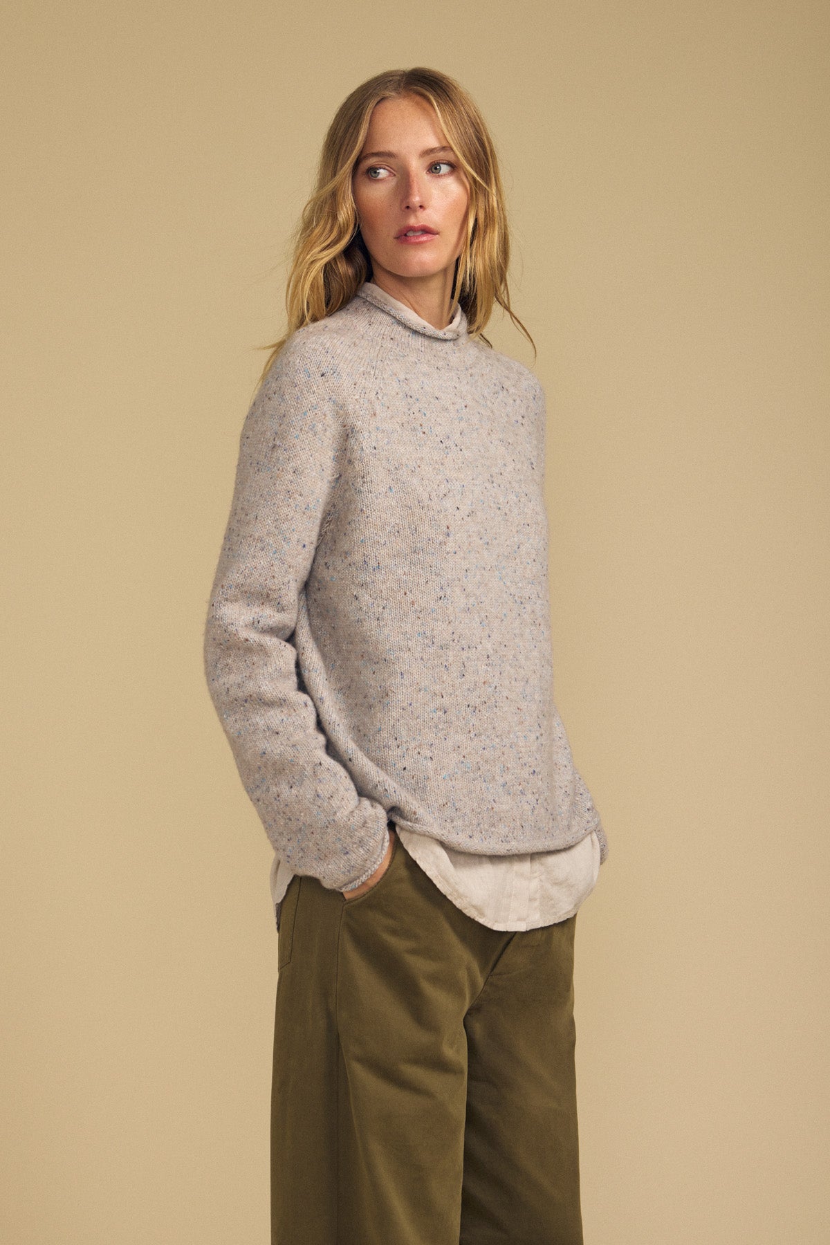 A woman wearing the JOELLE SWEATER by Velvet by Graham & Spencer—a speckled beige wool blend—over a light shirt and olive green pants, stands with her hands in her pockets against a plain beige background.
