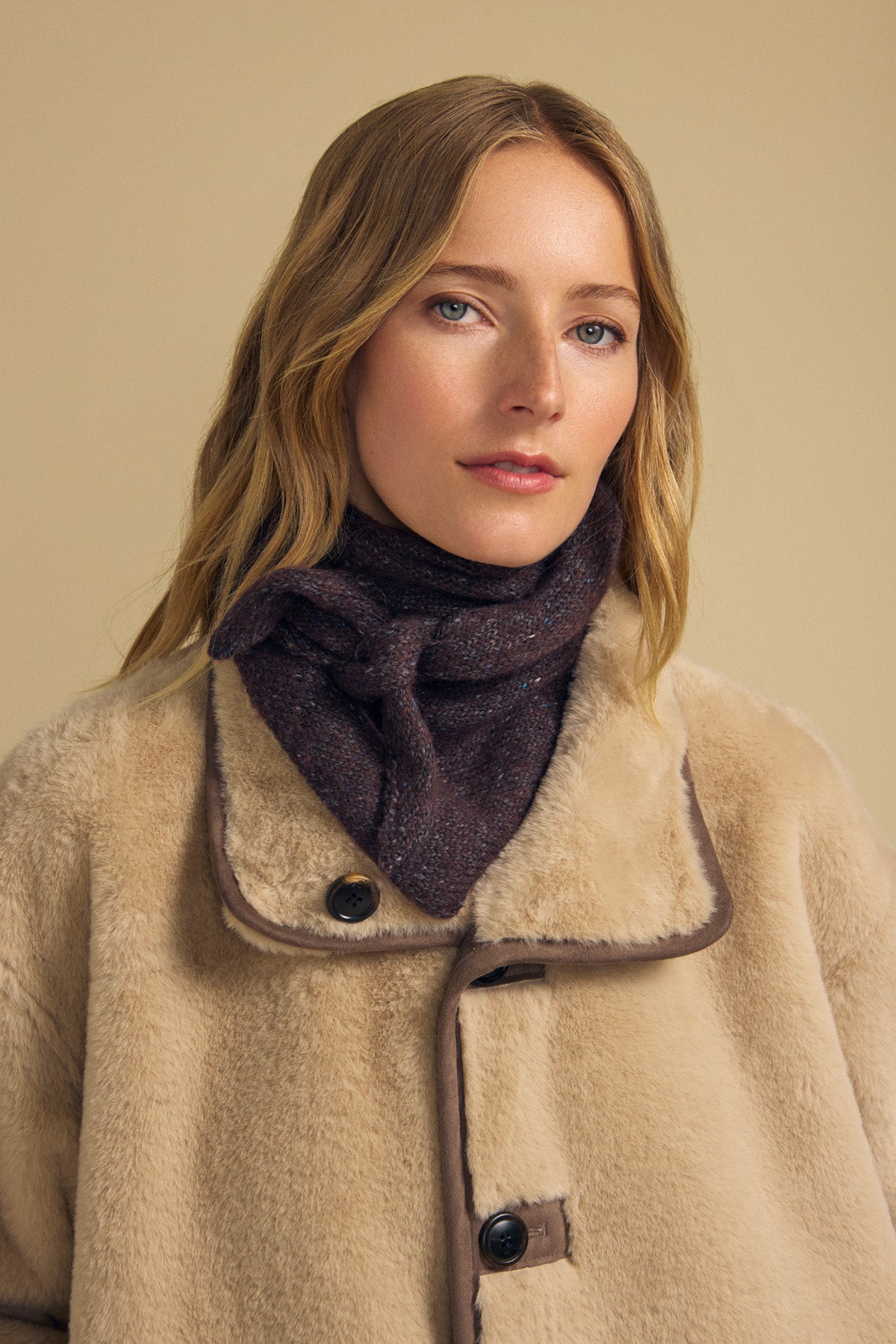 A woman with long blonde hair poses against a plain beige background, wearing a beige faux fur coat and the KARLA SCARF by Velvet by Graham & Spencer, a dark wool blend triangular scarf.