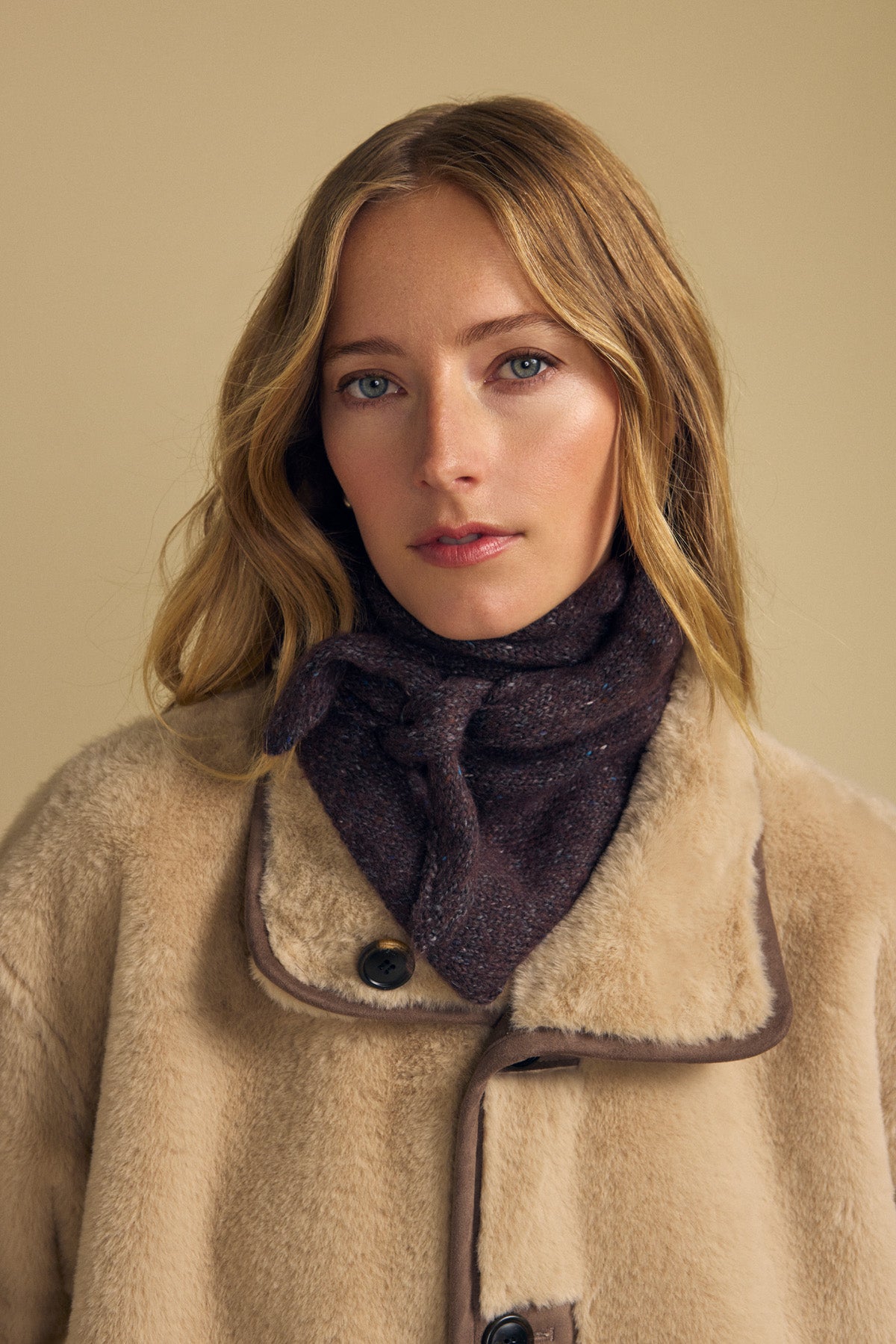 A woman with long blonde hair wears a beige shearling coat and the Velvet by Graham & Spencer KARLA SCARF—a dark wool-blend triangular scarf—as she looks at the camera against a plain light background, showcasing this perfect layering accessory for cooler days.