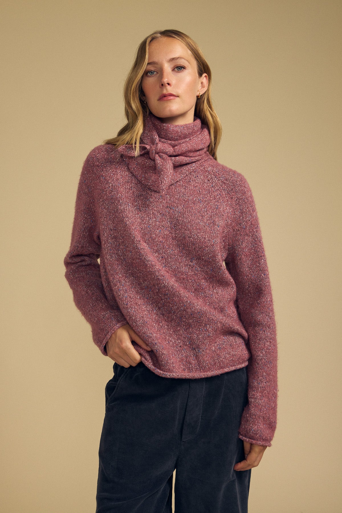 A woman styles the KARLA SCARF by Velvet by Graham & Spencer—a chic wool blend accessory—over a textured pink turtleneck sweater with a knotted collar and dark pants, standing against a beige background.