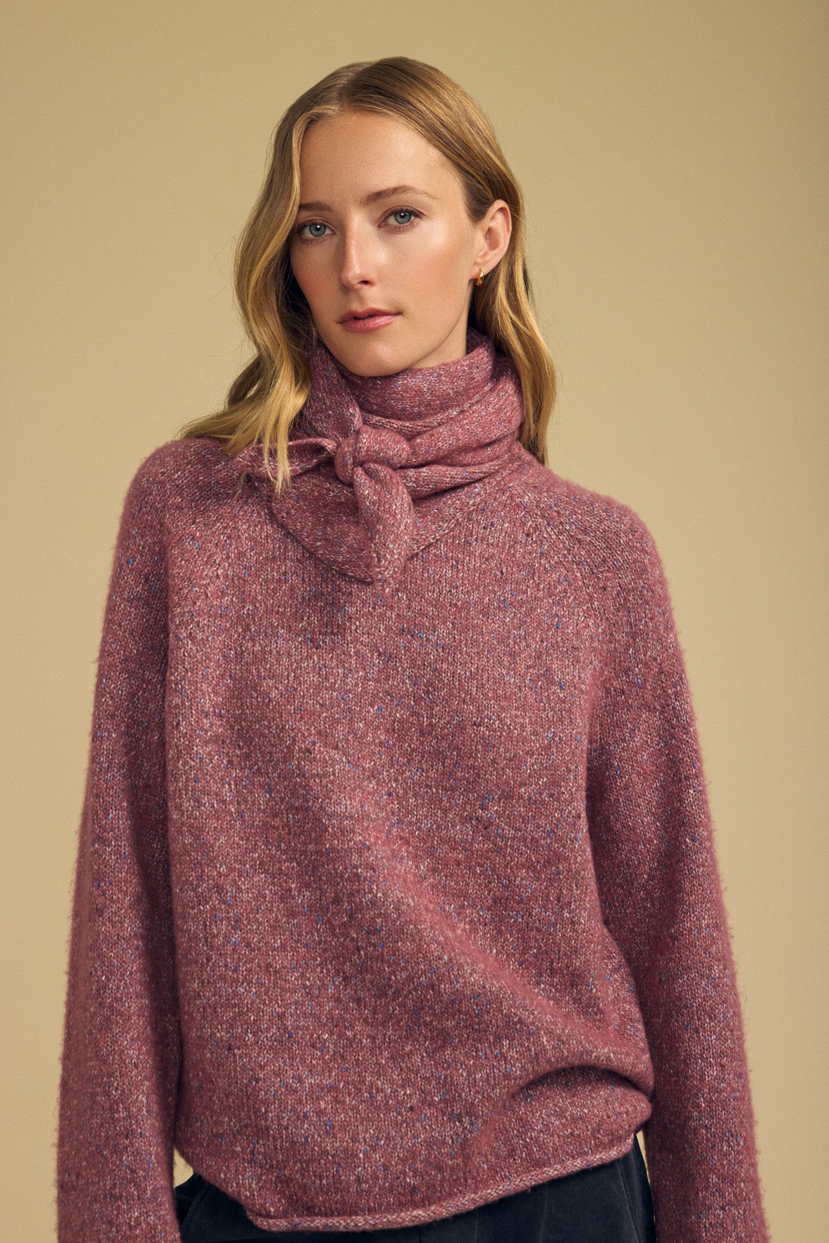 A woman stands against a plain beige background, styled with Velvet by Graham & Spencer’s KARLA SCARF—a pink wool blend accent—tied around her neck to complement her textured long-sleeve sweater.-42063771173057