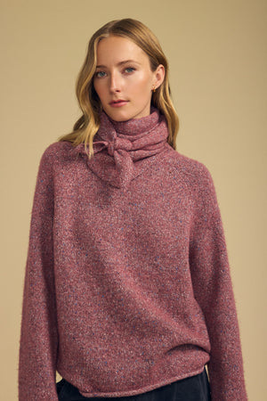 A woman stands against a plain beige background, styled with Velvet by Graham & Spencer’s KARLA SCARF—a pink wool blend accent—tied around her neck to complement her textured long-sleeve sweater.