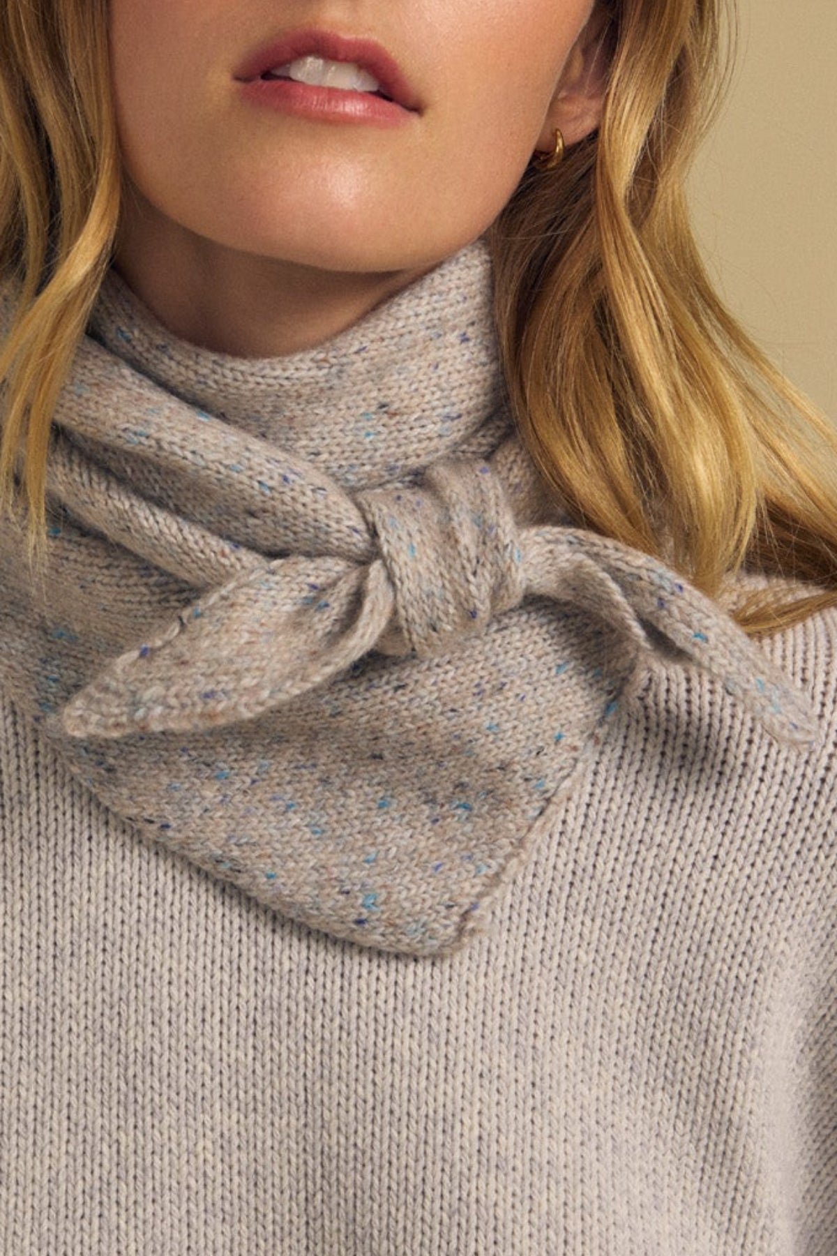 Close-up of a woman wearing a light gray speckled knit sweater and the Velvet by Graham & Spencer KARLA SCARF, a matching wool blend accessory elegantly knotted at the neck for cozy layering.-42063771238593