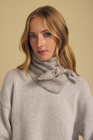 A woman with long blonde hair stands against a plain beige background, wearing a light grey knit wool blend sweater and the KARLA SCARF by Velvet by Graham & Spencer—a light grey speckled triangular scarf elegantly tied around her neck, making it the perfect layering accessory for understated elegance.