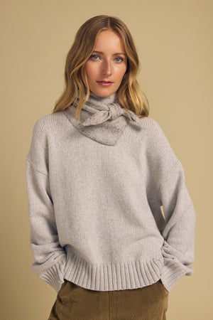 A woman with long blonde hair stands against a beige background, wearing a light gray sweater, tan pants, and the Velvet by Graham & Spencer KARLA SCARF—chicly tied around her neck.