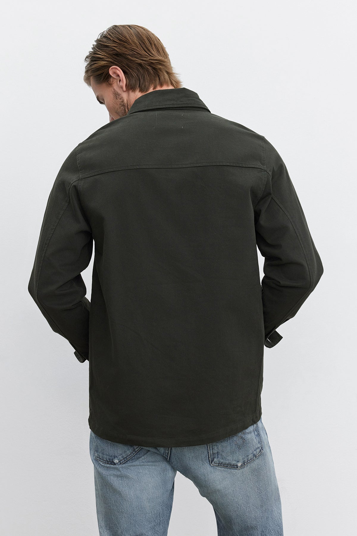   A person with light brown hair facing away, wearing the BARCLAY CHORE JACKET by Velvet by Graham & Spencer, crafted from mid-weight cotton lyocell twill in dark green, paired with blue jeans, stands against a plain light background. 